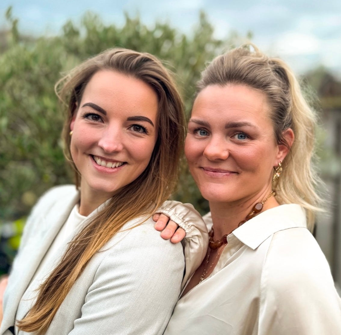 Twee vrouwen lachen naast elkaar in een natuurlijke buitenomgeving, portret van de oprichters van NU-MI.
