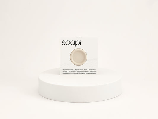 Witte Soapi magnetische zeep houder in verpakking gepresenteerd op ronde witte display tegen lichte beige achtergrond.