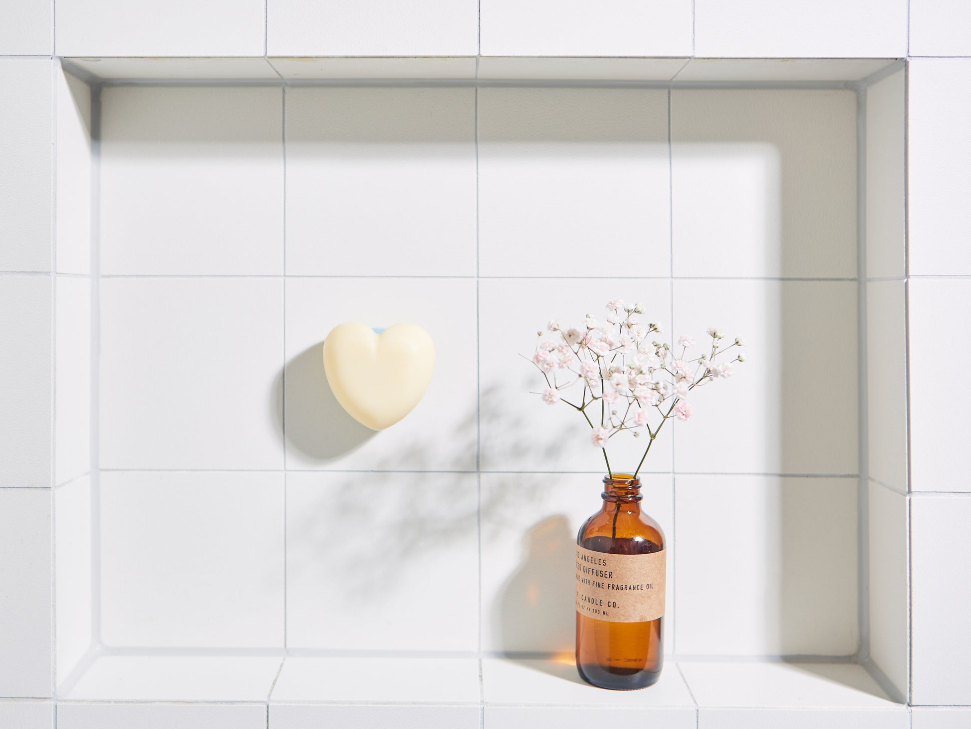 Hartvormige NU-MI zeepbar op een witte tegelnis naast een glazen flesje met droogbloemen.