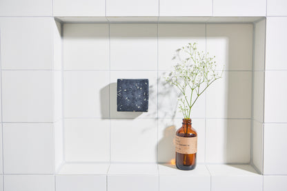 Zwarte handgemaakte zeep aan magnetische houder in betegelde badkamer nis met glazen flesje en bloemen.