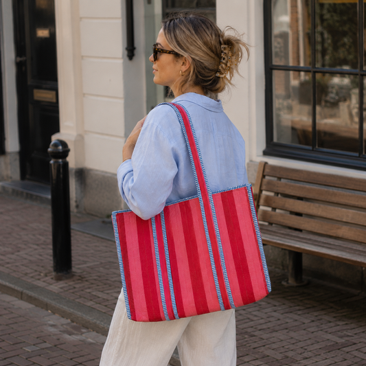 Vrouw draagt een grote NU-MÍ tote bag met roze en rode strepen in een Nederlandse stadsomgeving.